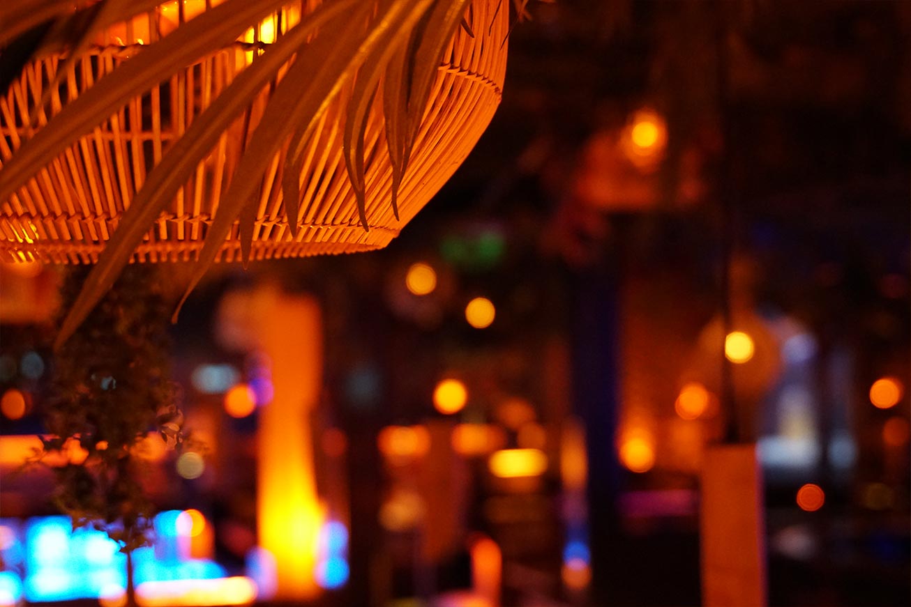 Geflochtener Rattan-Lampenschirm mit warmem Licht vor unscharf leuchtendem Bar-Hintergrund.